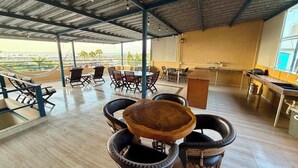 Uma área de estar coberta ao ar livre com mesas e cadeiras, uma mesa de madeira no centro e vista para o mar.