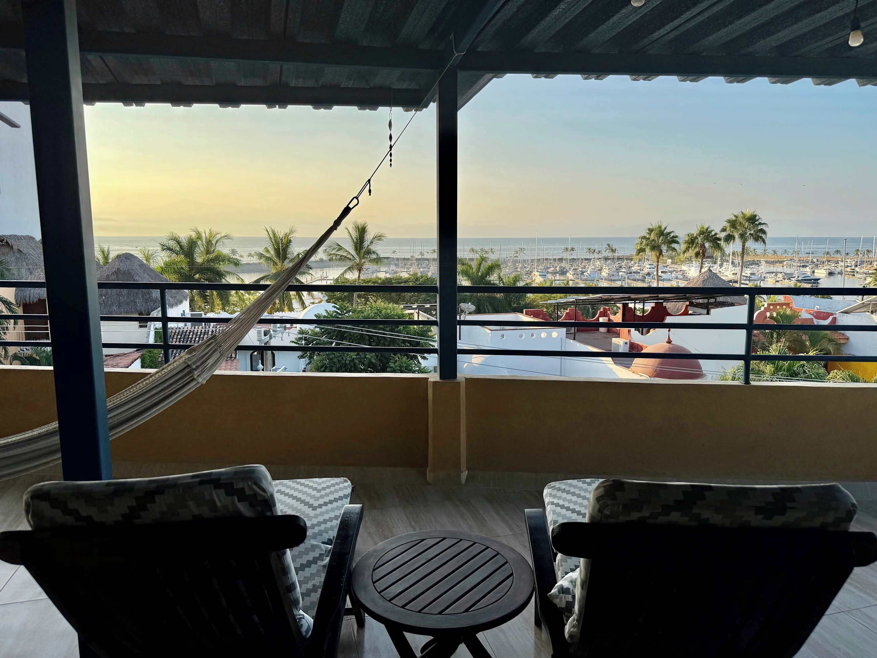 A balcony with a hammock, two chairs, and a small table. A view of a marina with boats and palm trees is visible.