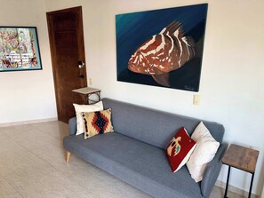 A living room with a grey sofa, two patterned cushions, a small wooden table, and a large fish painting on the wall. - Rooftop Patio, Marina Views––1 Block to Beach (La Cruz de Huanacaxtle)