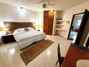 A hotel room with a bed, bedside lamps, a ceiling fan, a wooden wardrobe, a small shelf with books, and a door leading to another room. - Rooftop Patio, Marina Views––1 Block to Beach (La Cruz de Huanacaxtle)