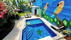 Piscina com um mural de araras e uma tartaruga, rodeada por espreguiçadeiras e um pequeno edifício.