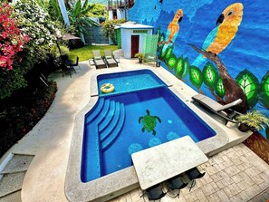 Ein Swimmingpool mit einem Wandgemälde von Papageien und einer Schildkröte, umgeben von Liegestühlen und einem kleinen Gebäude.