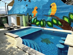 Ein Swimmingpool mit Wandgemälde von Papageien und Bäumen, Liegestühlen und einem kleinen Gebäude.