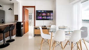 Een moderne woonkamer met een glazen eettafel, witte stoelen, een flatscreen-televisie aan de muur en een keuken met een bar en zitplaatsen.