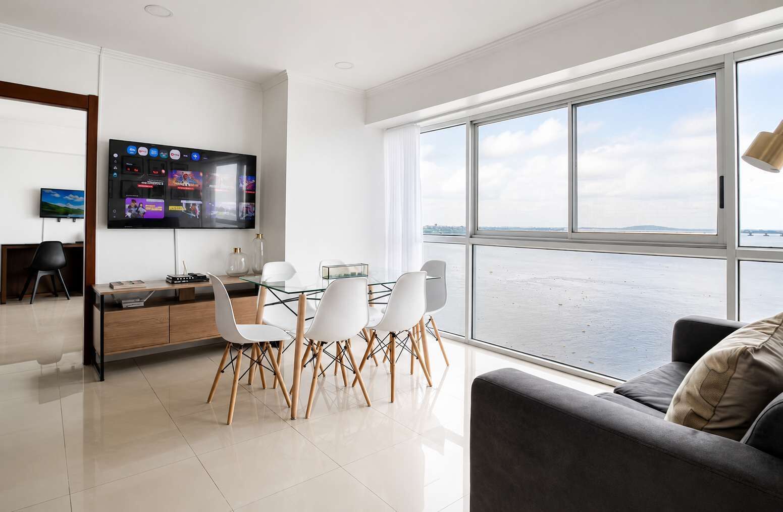 A modern living room with a large window, a flat-screen TV, and a dining area with white chairs.