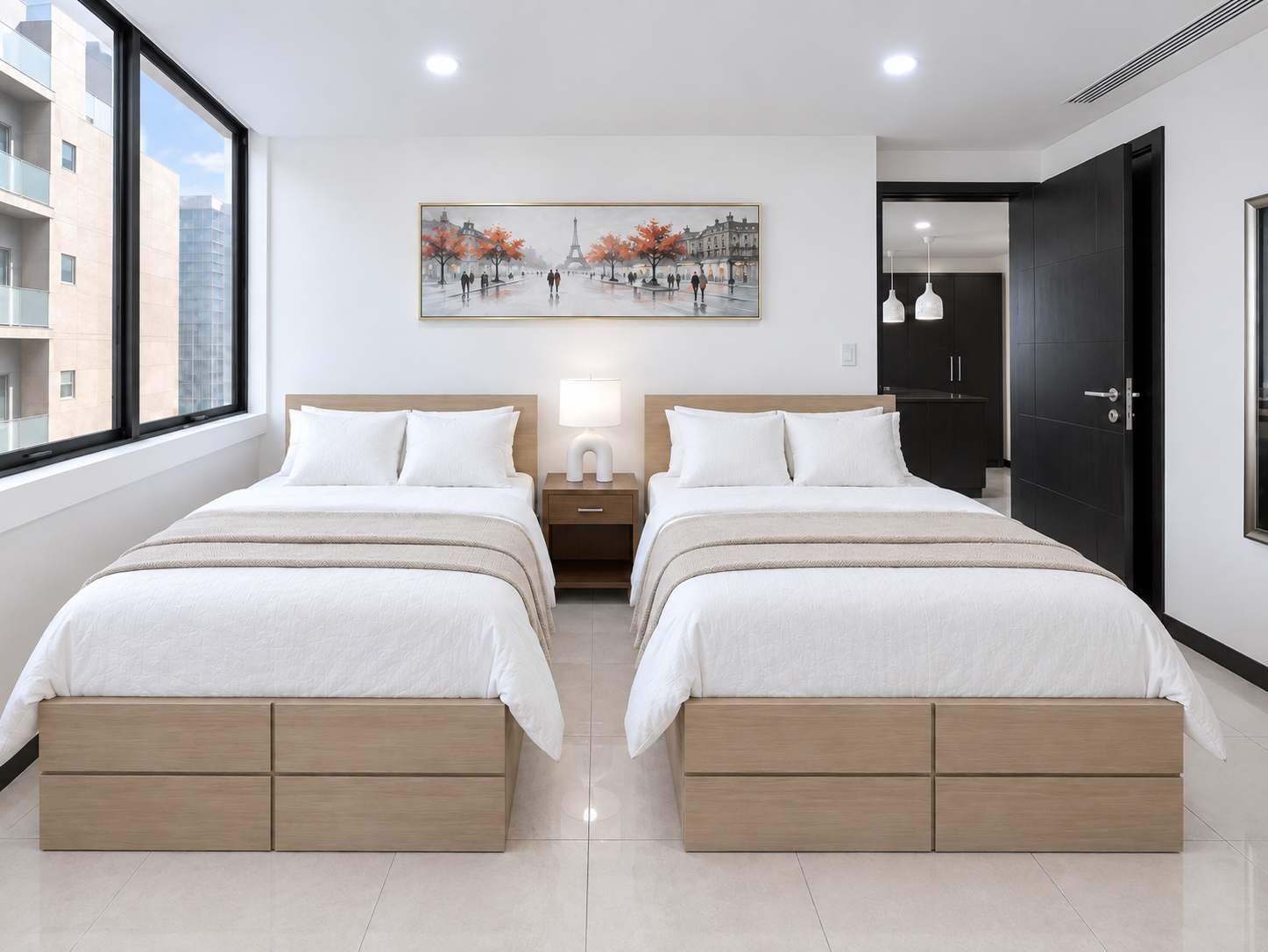 Een moderne hotelkamer met twee bedden, een nachtkastje met een lamp, een schilderij van een stadsgezicht en uitzicht op gebouwen.