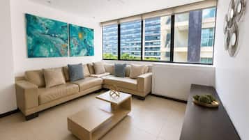 Un salon moderne avec un canapé d’angle beige, une table basse et une grande fenêtre donnant sur des immeubles.