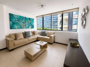 Un salon moderne avec un canapé d’angle beige, une table basse et une grande fenêtre donnant sur des immeubles.