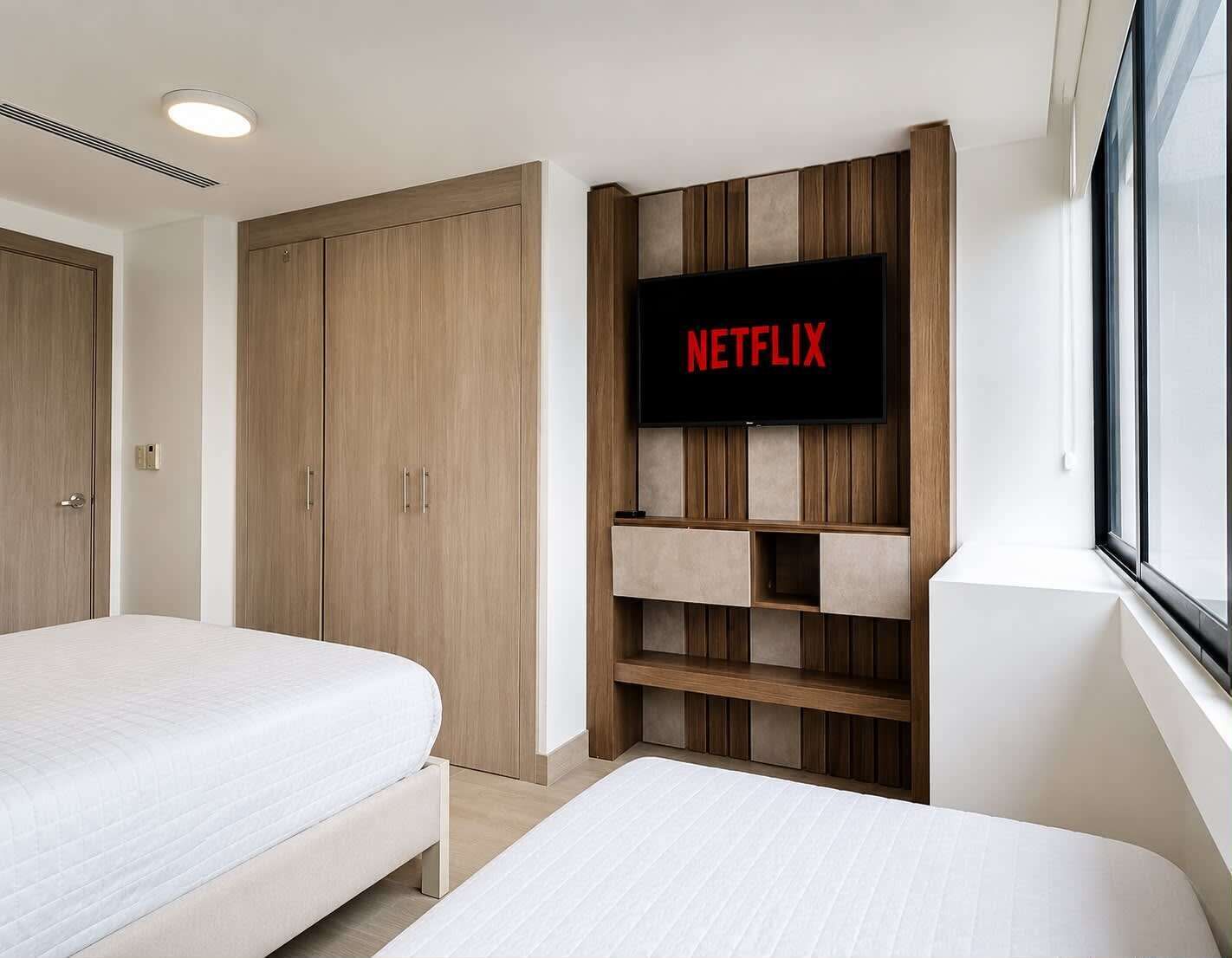 A bedroom with two beds, a wardrobe, a television mounted on a wooden panel wall, and a window.