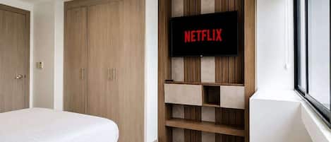 A bedroom with two beds, a wardrobe, a television mounted on a wooden panel wall, and a window.