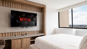 Habitación de hotel moderna con un televisor de pantalla plana grande fijado a una pared de paneles de madera, una cama con un edredón blanco acolchado y vistas de un paisaje urbano a través de un amplio ventanal.