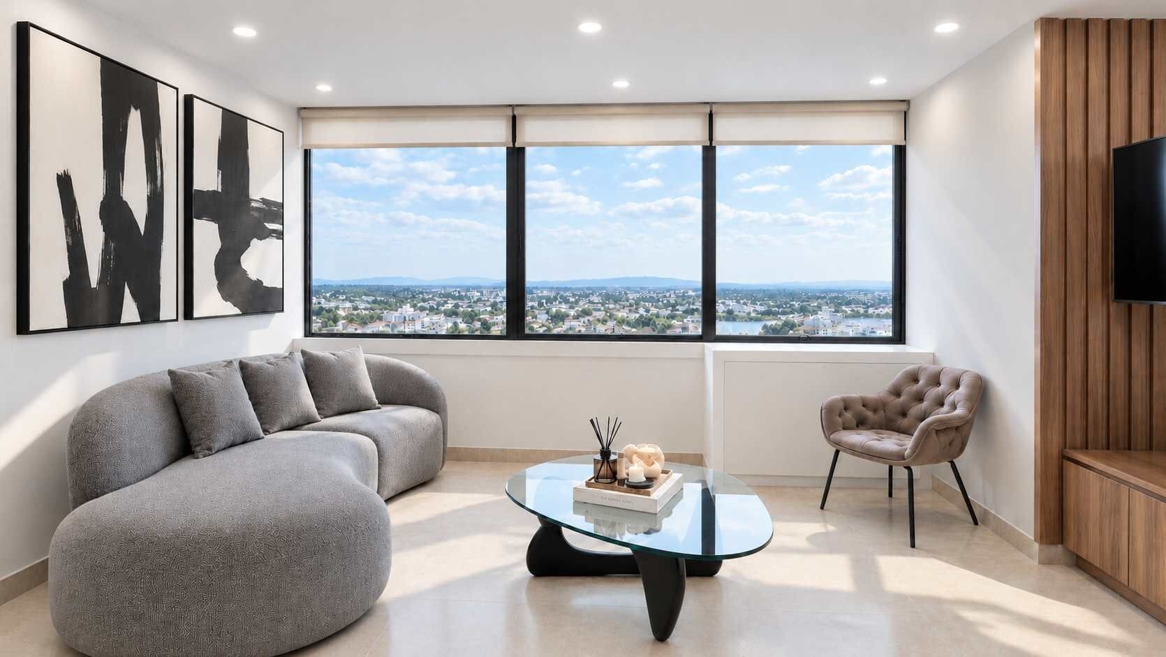 Ein modernes Wohnzimmer mit einem grauen Sofa, einem Glastisch und Blick auf eine Stadtkulisse durch große Fenster.