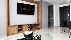 Una sala de estar moderna con un televisor de pantalla plana montado en una pared de madera, una mesa de centro de vidrio con un cuenco decorativo y un sofá de color oscuro.