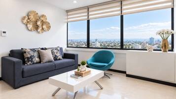 A modern living room with a sofa, a coffee table, a blue chair, and a city view through large windows.