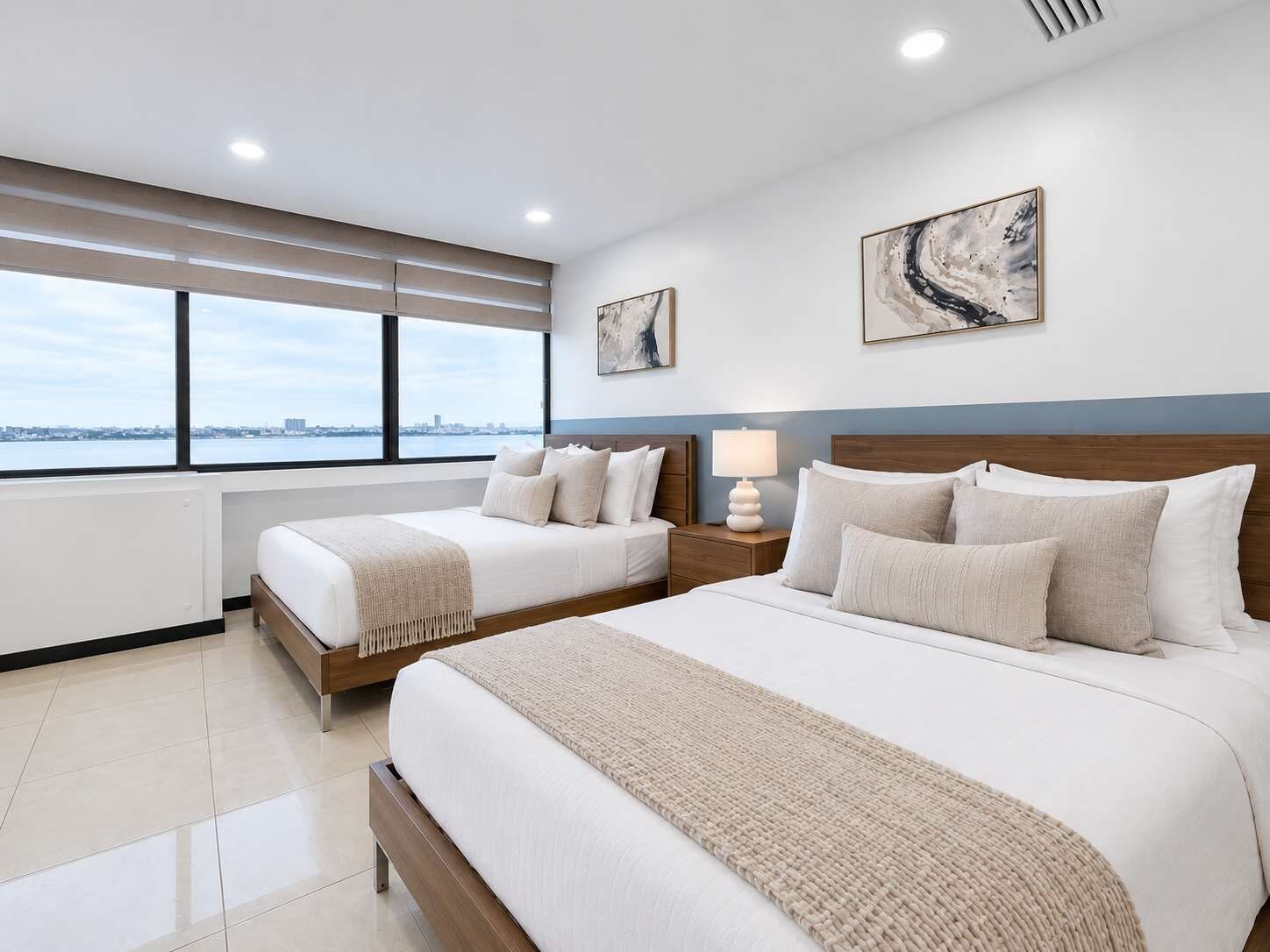 A hotel room with two beds, a large window with a city view, and artwork on the walls.