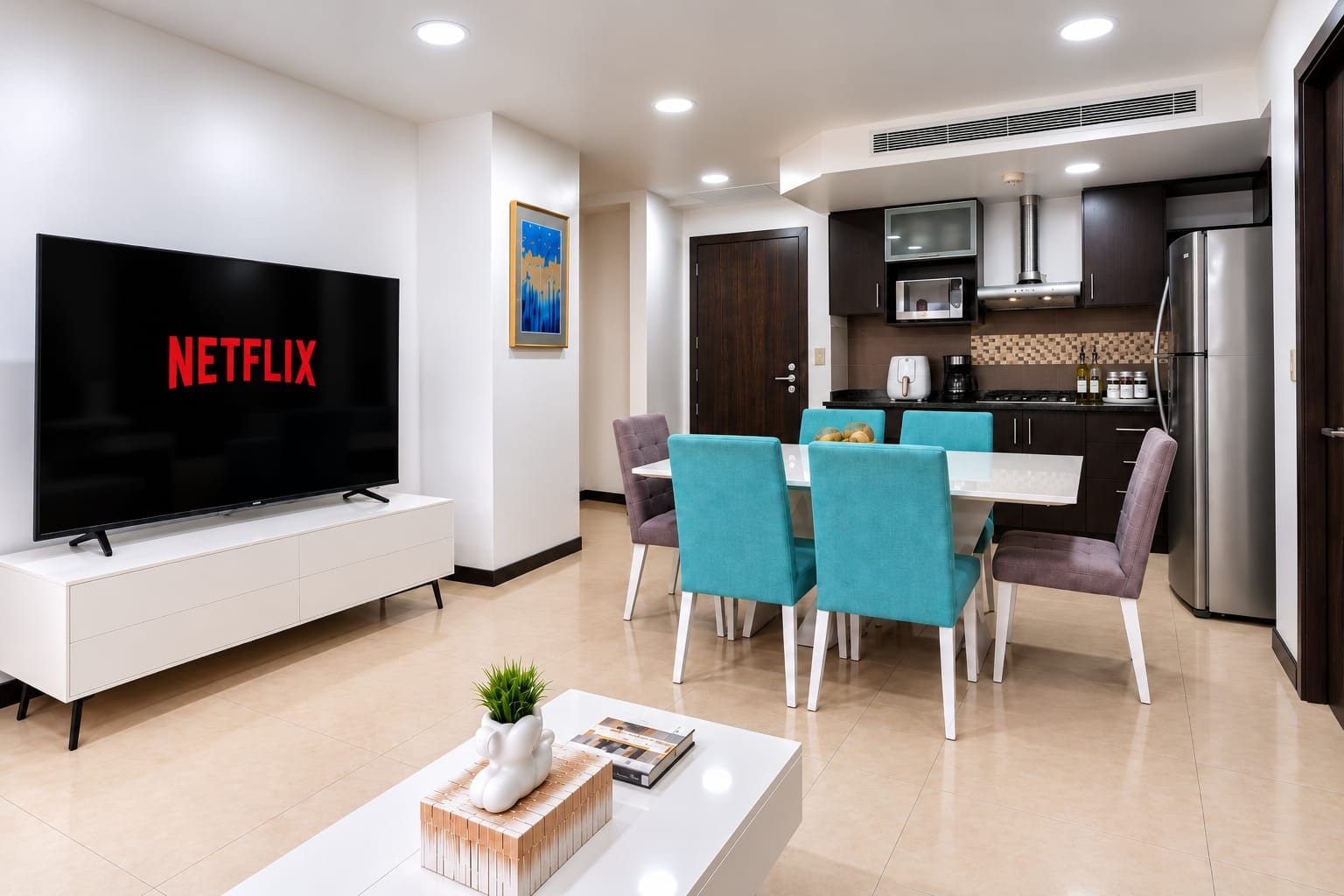 A modern living room with a flat-screen TV, a dining area with teal chairs, and a kitchen with dark cabinets and stainless steel appliances.