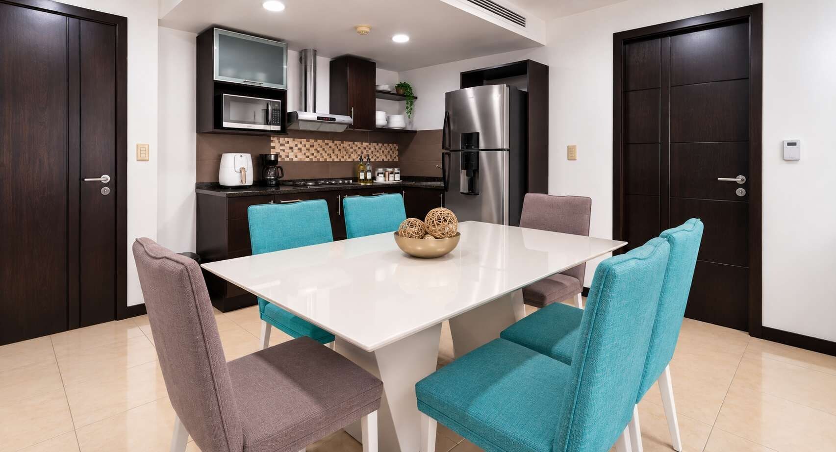 A modern kitchen with dark wood cabinets, a stainless steel refrigerator, and a white dining table with teal chairs.