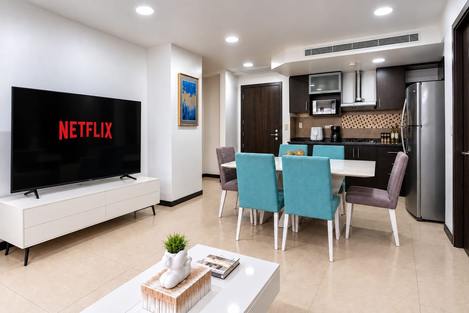 A modern living room with a flat-screen TV displaying 'NETFLIX,' a dining area with teal chairs, and a kitchen with dark cabinets and stainless steel appliances.