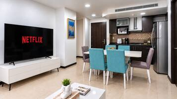 A modern living room with a flat-screen TV displaying 'NETFLIX,' a dining area with teal chairs, and a kitchen with dark cabinets and stainless steel appliances.