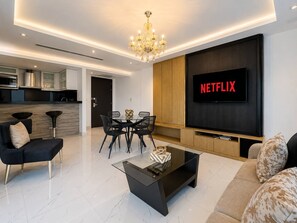 Moderni olohuone, jossa on litteänäyttöinen televisio, jonka näytöllä näkyy teksti »NETFLIX», ruokailutila lasipöydällä ja -tuoleilla sekä musta sohva kuviollisilla koristetyynyillä.