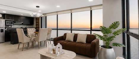 A modern living room with a sofa, chairs, a coffee table, and a large window with a view of the sunset.