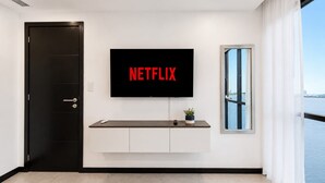 Habitación moderna con una gran televisión de pantalla plana montada en la pared que muestra 'NETFLIX' en letras rojas, una planta pequeña sobre una consola y vista al agua a través de una ventana.