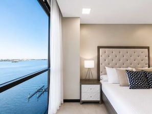 A hotel room with a bed, nightstand, lamp, and a view of the water. - The PANORAMA | River View Suite | Puerto Santa Ana (Guayaquil)
