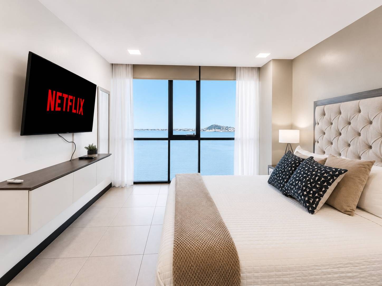 Une chambre d’hôtel avec un lit, un téléviseur affichant « NETFLIX », une vue sur la mer et une fenêtre avec des rideaux.