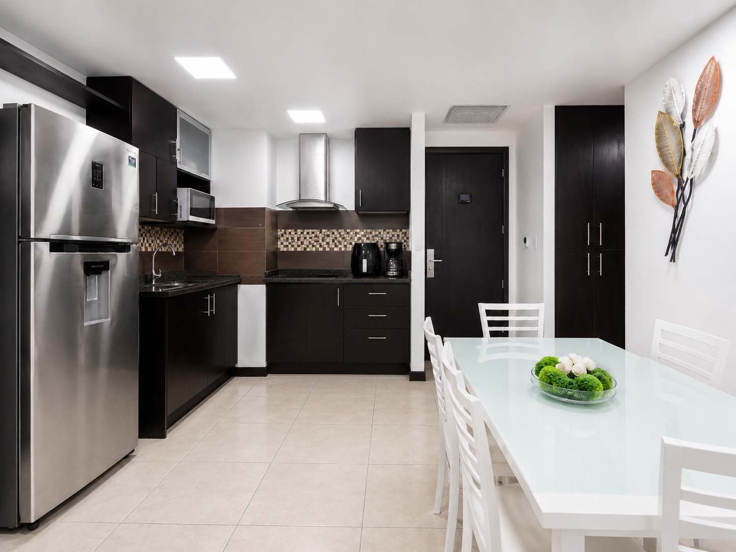 Una cocina moderna con un refrigerador de acero inoxidable, un microondas y una cafetera. Hay una mesa de comedor con sillas y un tazón de verduras verdes.
