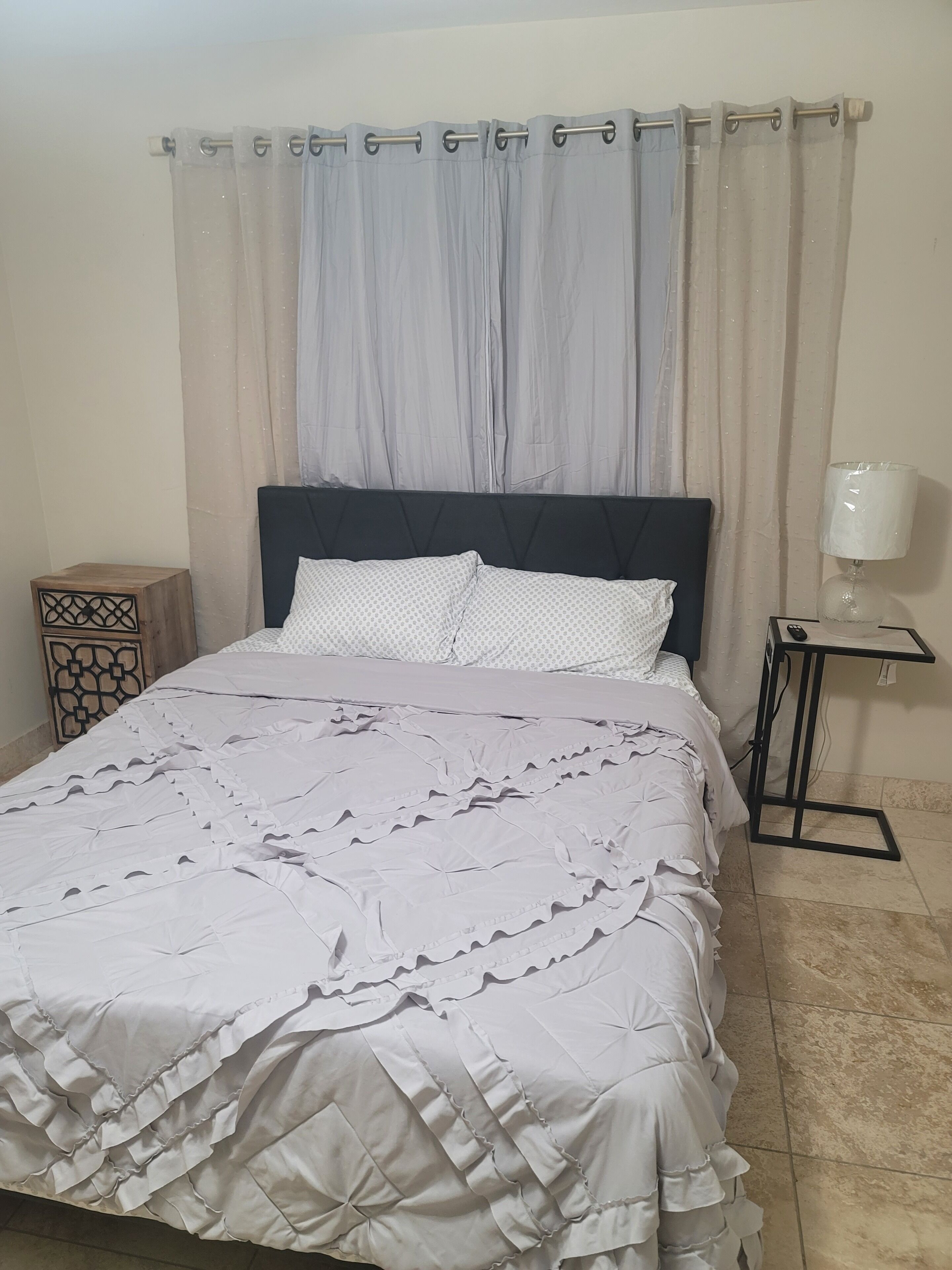 A bed with a dark headboard, a white patterned bedspread, and a decorative pillow. There is a nightstand with a lamp and a small table with a remote control.