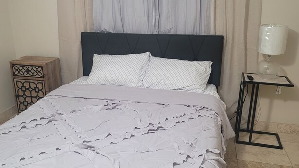 A bed with a dark headboard, a white patterned bedspread, and a decorative pillow. There is a nightstand with a lamp and a small table with a remote control. - Marvelous 3-bedroom house with AC big Pool and Jaccuzi very quiet in Lehigh Acre (Lehigh Acres)