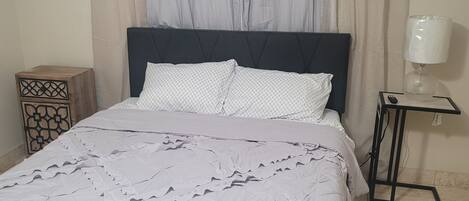 A bed with a dark headboard, a white patterned bedspread, and a decorative pillow. There is a nightstand with a lamp and a small table with a remote control.