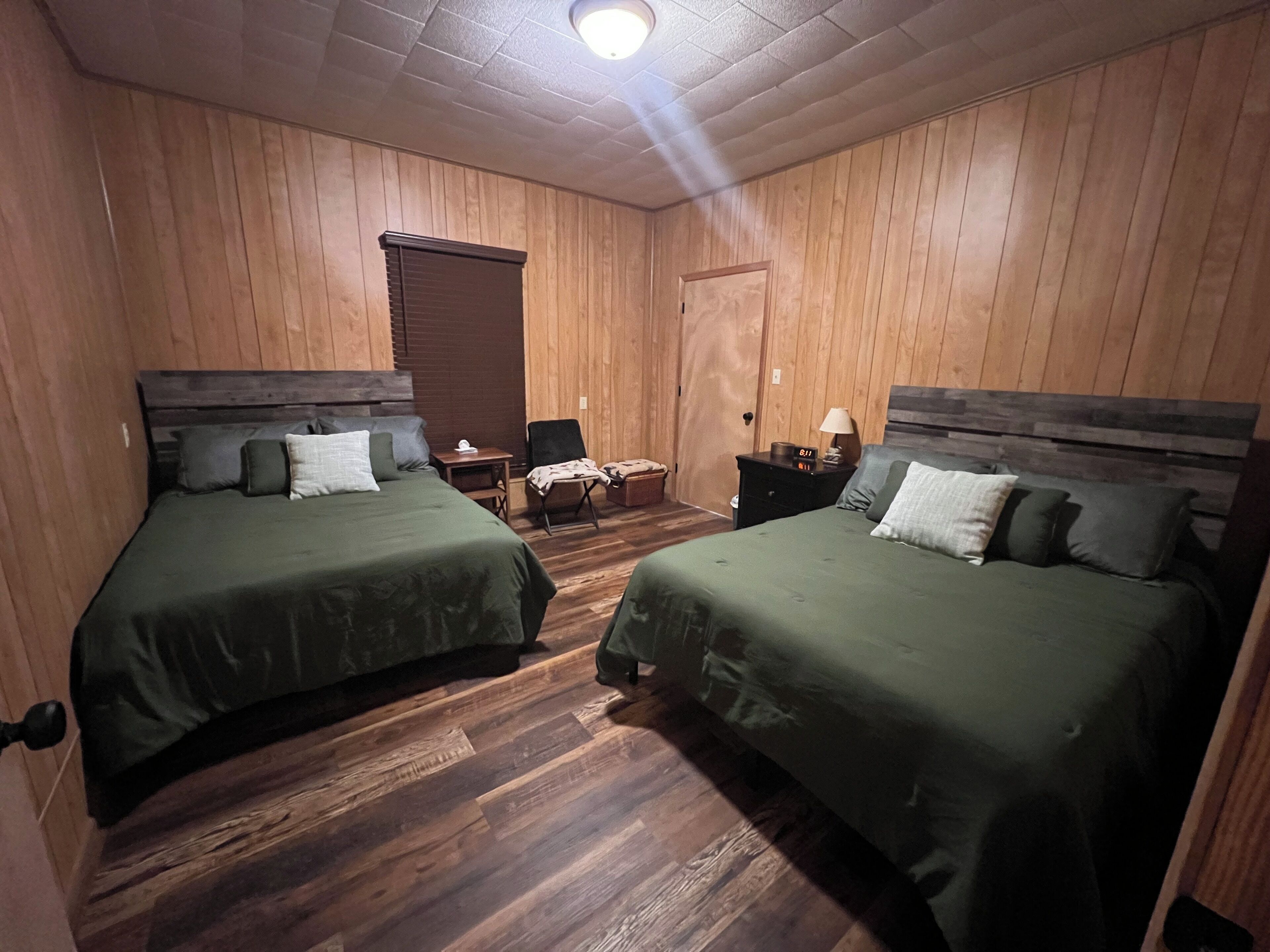A room with two beds, wooden flooring, a chair, a nightstand with a lamp, and a door.