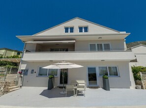A two-story house with a balcony, a patio with outdoor furniture, and a clear blue sky. - Two Bedroom Apartment with Terrace (Mlini)