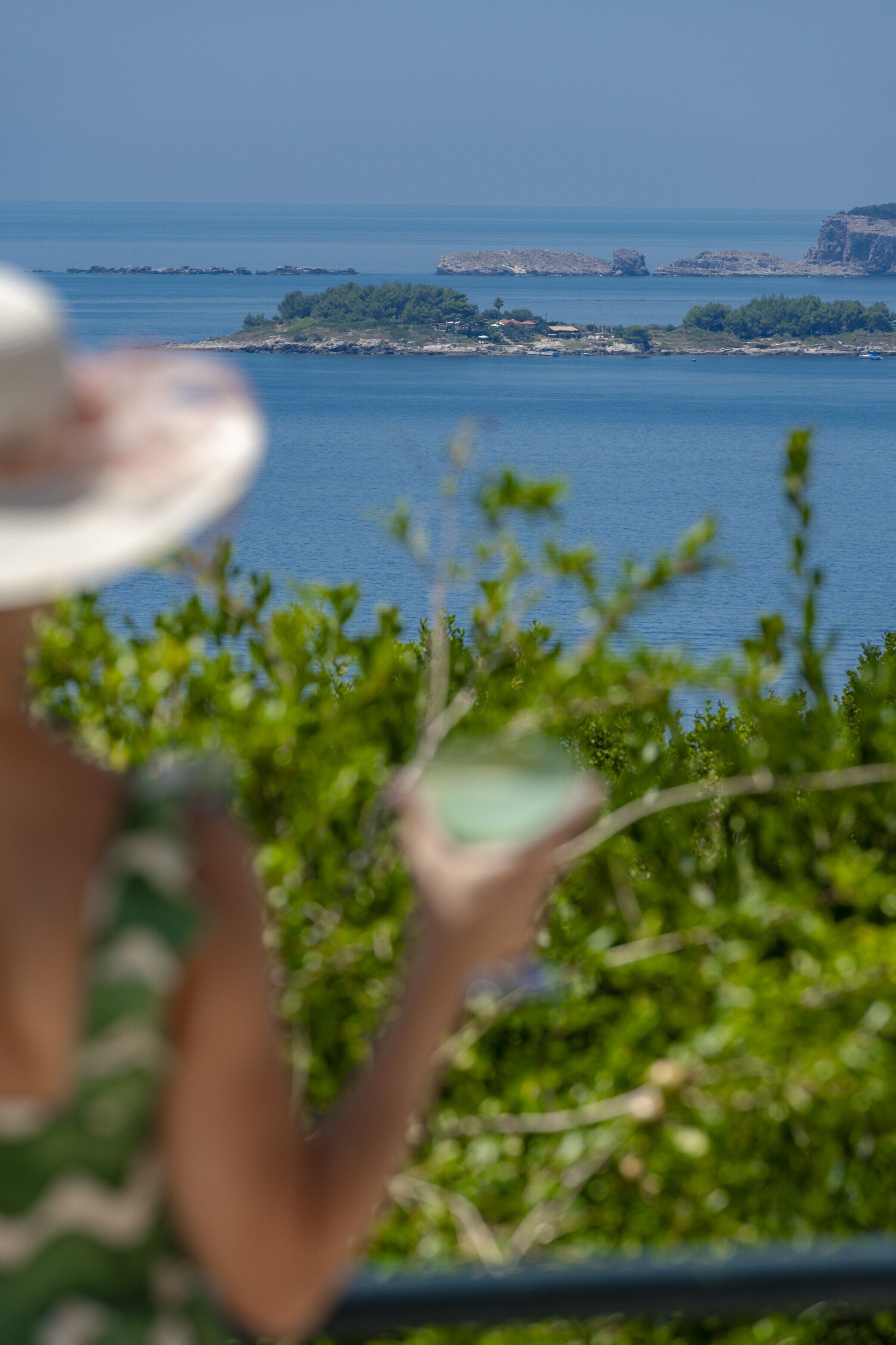 A person holding a drink, overlooking a coastal landscape with buildings and greenery.