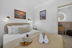 Ein modernes Hotelzimmer mit einem Bett, zwei Wandlampen, einem Gemälde, einem Badezimmer mit Spiegel und einem Tablett mit Handtüchern und einem Croissant.