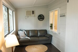 Ein Zimmer mit einer Ledersofa, einem runden Webteppich, einer Wandklimaanlage und einem Fenster mit Blick ins Grüne.