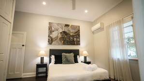 A bedroom with a large bed, two bedside tables with lamps, a ceiling fan, an air conditioning unit, and a window with curtains. - The Foxman Cottage (Broke)