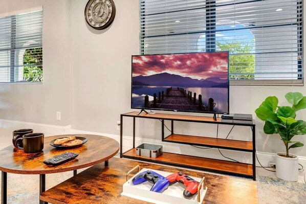 Ein Wohnzimmer mit einem Fernseher auf einem schwarzen Standfuß, einem Couchtisch aus Holz mit Fernbedienung und Snacks sowie einer Pflanze rechts.