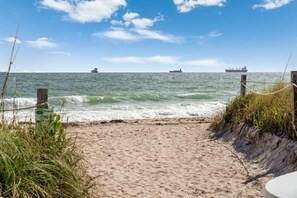 Ein sandiger Strand mit einer Holzzaun und einem Schild, das zum Meer führt, wo Schiffe sichtbar sind.