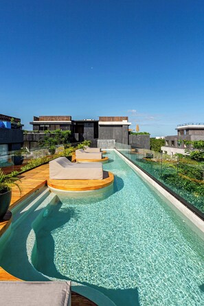A rooftop infinity pool with clear blue water, wooden deck seating, and a view of modern buildings. - Modern 1BR Jungle View Retreat in Humana | Rooftop (Tulum)