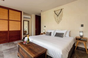 Ein Hotelzimmer mit einem großen Bett, Nachttischen, einem Holzbeistelltisch, einer wandmontierten Makramee-Dekoration und einem Holztrenner.