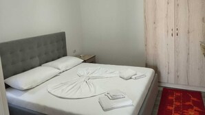 En säng för en person med en grå, klädd sänggavel, ett sängbord med en lampa och en garderob i trä med två dörrar.