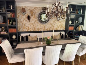 Een eetruimte met een lange tafel, gestreepte stoelen, een decoratieve wand met een spiegel, en een kroonluchter.