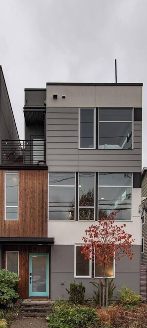 Ein mehrgeschossiges Wohngebäude mit modernem architektonischen Design, das Holz und Metall kombiniert, große Fenster besitzt und einen Balkon im oberen Geschoss zeigt.