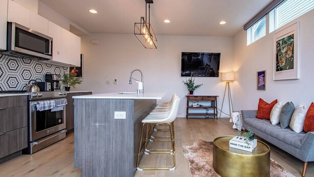 A modern kitchen with a central island, bar stools, a flat-screen TV, and a grey sofa with red cushions. - Modern 3 Bedroom Townhouse w/ Private Rooftop (Seattle)