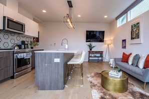 Eine moderne Küche mit Kochinsel, Barhocker, einem Flachbildfernseher und einem grauen Sofa mit roten Kissen.