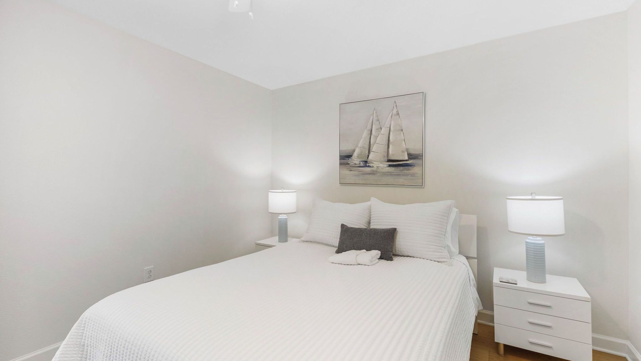 A bedroom with a bed, two bedside tables, a nightstand, a painting of a sailboat, and a lamp on each table.