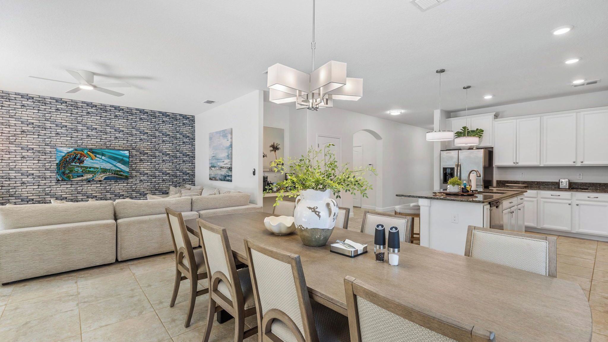 A modern kitchen with white cabinets, a dining area with a wooden table and chairs, and a brick wall with a painting.