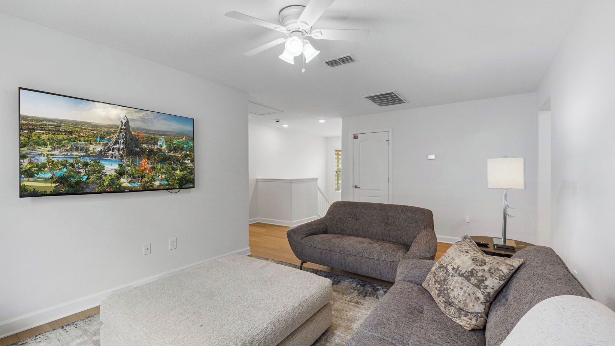 A living room with a flat-screen TV, a gray sofa, a patterned throw pillow, a white armchair, and a ceiling fan.